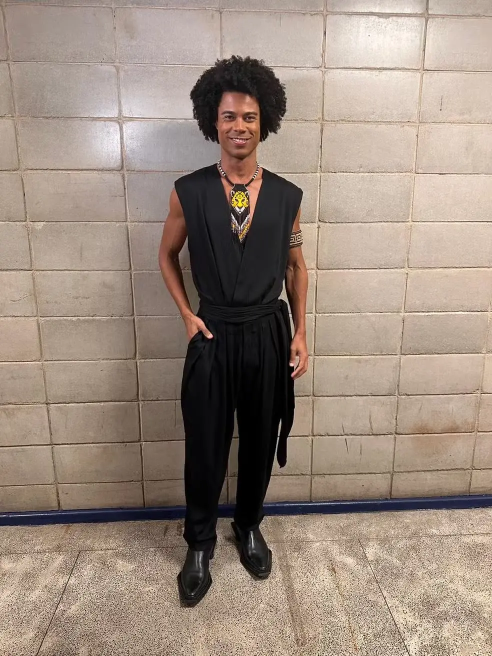 Breno é um homem jovem negro de cabelo black power. Na foto, ele usa um macacão preto com um colar de leão e um acessório no braço.
