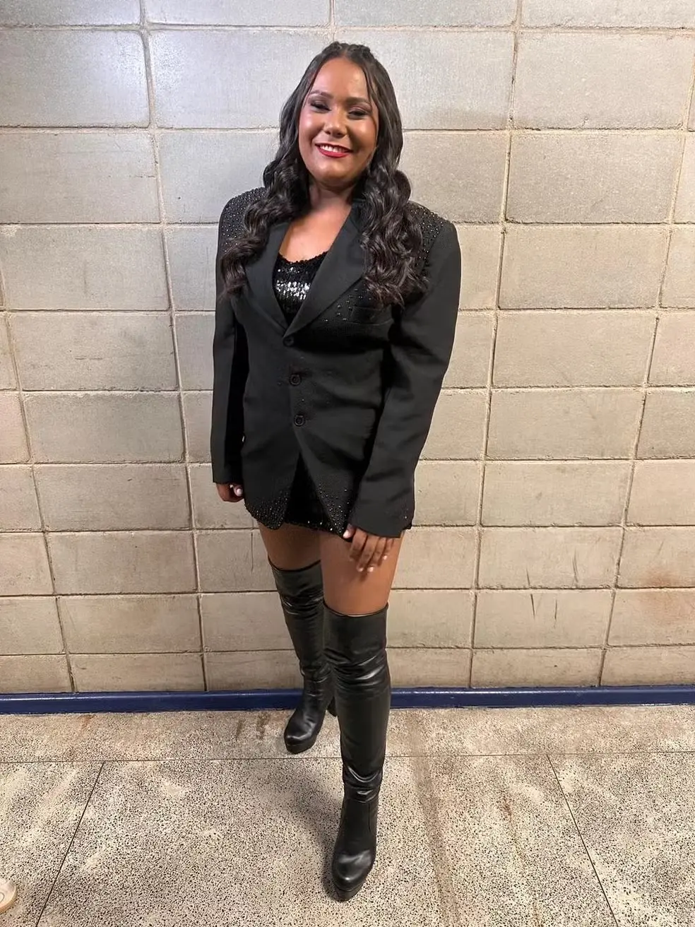 Chaiany é uma mulher jovem negra de cabelo castanho escuro ondulado. Na foto, ela está de terno preto e bota da mesma cor.