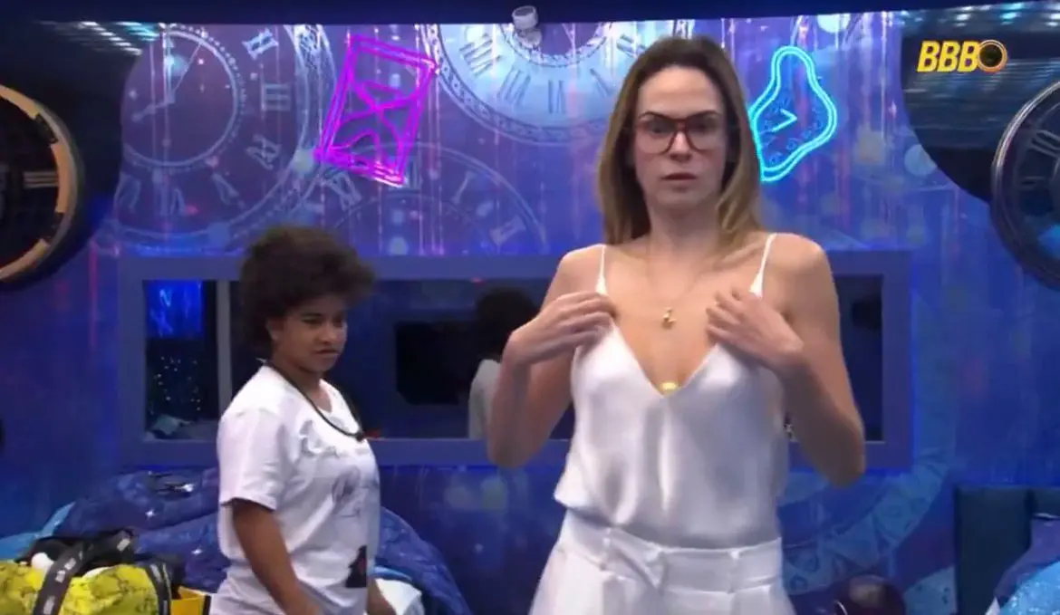 Ana Paula experimenta um top de cetim branco com alças finas e decote em V, peça escolhida para a final do BBB 26, enquanto Milena observa ao fundo em um quarto decorado com relógios.