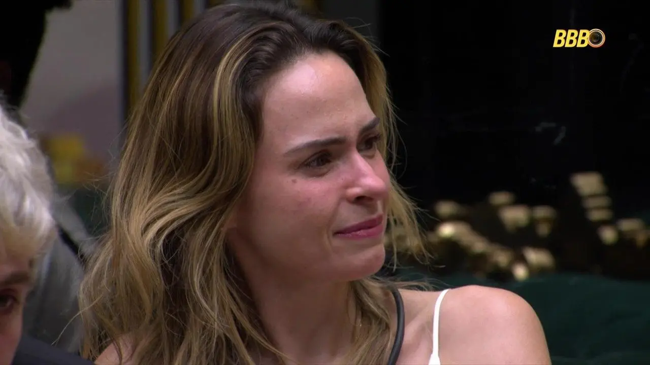 Ana Paula Renault sentada em ambiente interno do BBB 26, vista de perfil, com cabelo solto, vestindo blusa de alça; logotipo do programa aparece no canto superior da imagem.