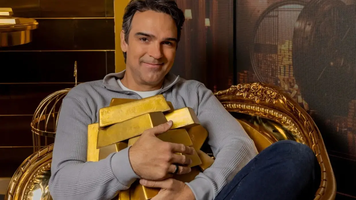 Tadeu Schmidt é um homem de meia idade, branco, de cabelo preto. Na foto, ele está de moletom cinza sentado em uma poltrona dourada abraçando barras de ouro. Imagem usada para informar que prêmio do BBB 26 aumenta de valor.