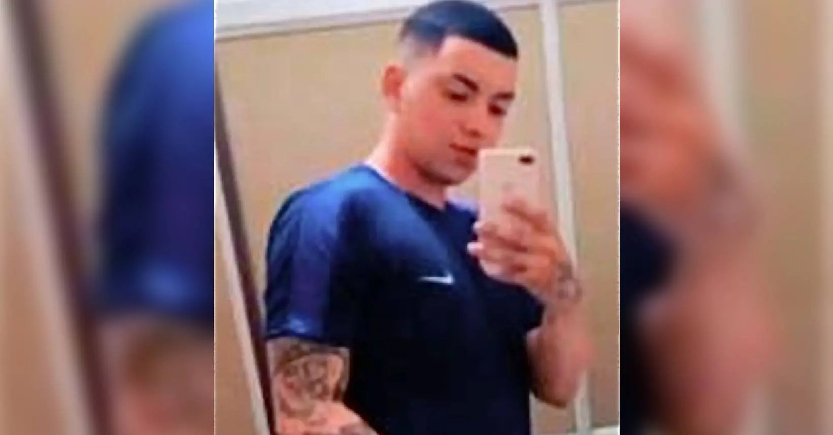 Homem tirando selfie em espelho, usando camisa azul, com cabelo curto e tatuagens no braço, em ambiente interno.