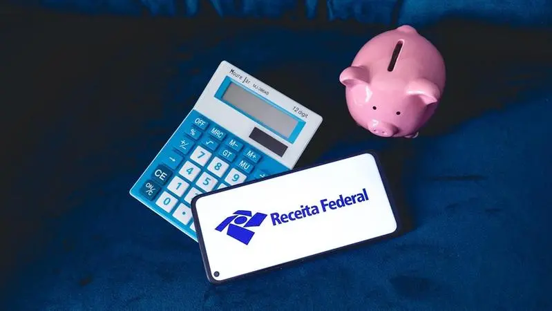 Imagem de uma calculadora, um cofrinho em formato de porquinho rosa e um celular que tem escrito Receita Federal na tela.