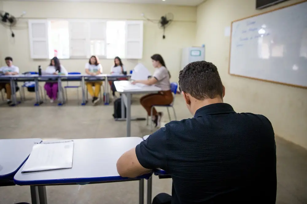 pessoa de costas, vestindo camisa escura, sentada em uma sala de aula.