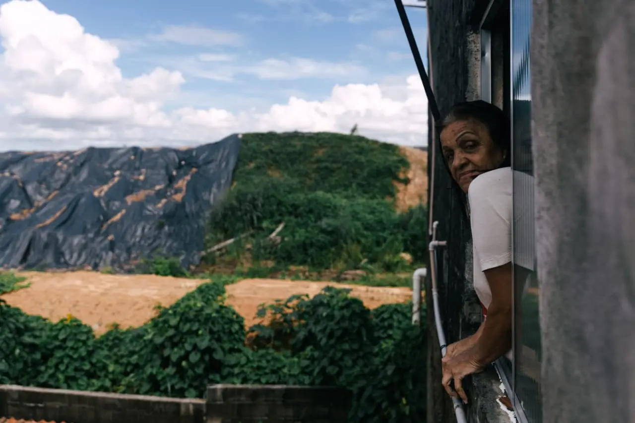 Mulher idosa olha pela janela de sua casa para um terreno coberto por lonas pretas, em um cenário de contraste entre a moradia e o local de obras.