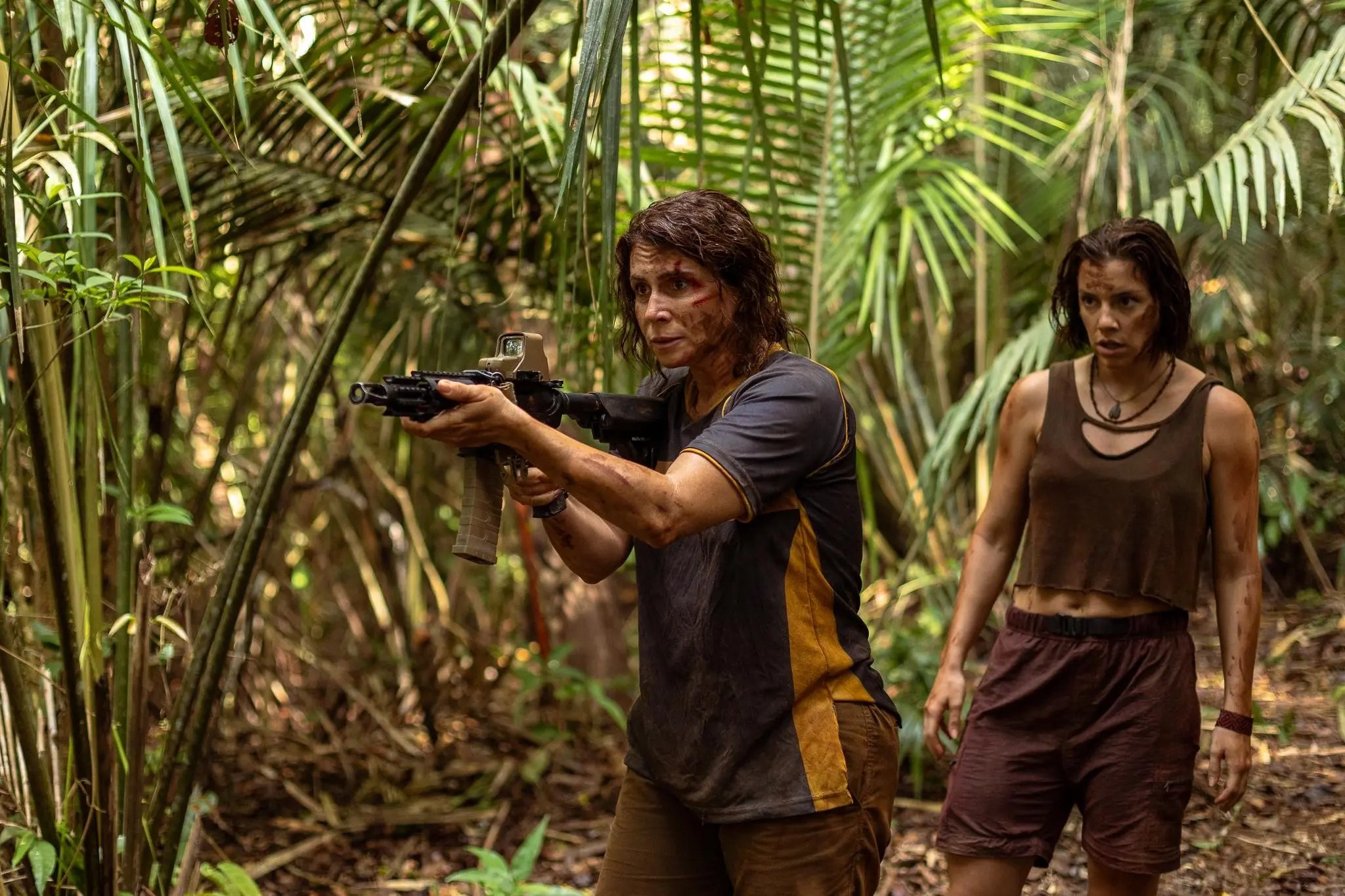 Duas pessoas em uma área de floresta densa; a pessoa em primeiro plano aponta uma arma enquanto caminha entre a vegetação, e outra pessoa segue logo atrás.