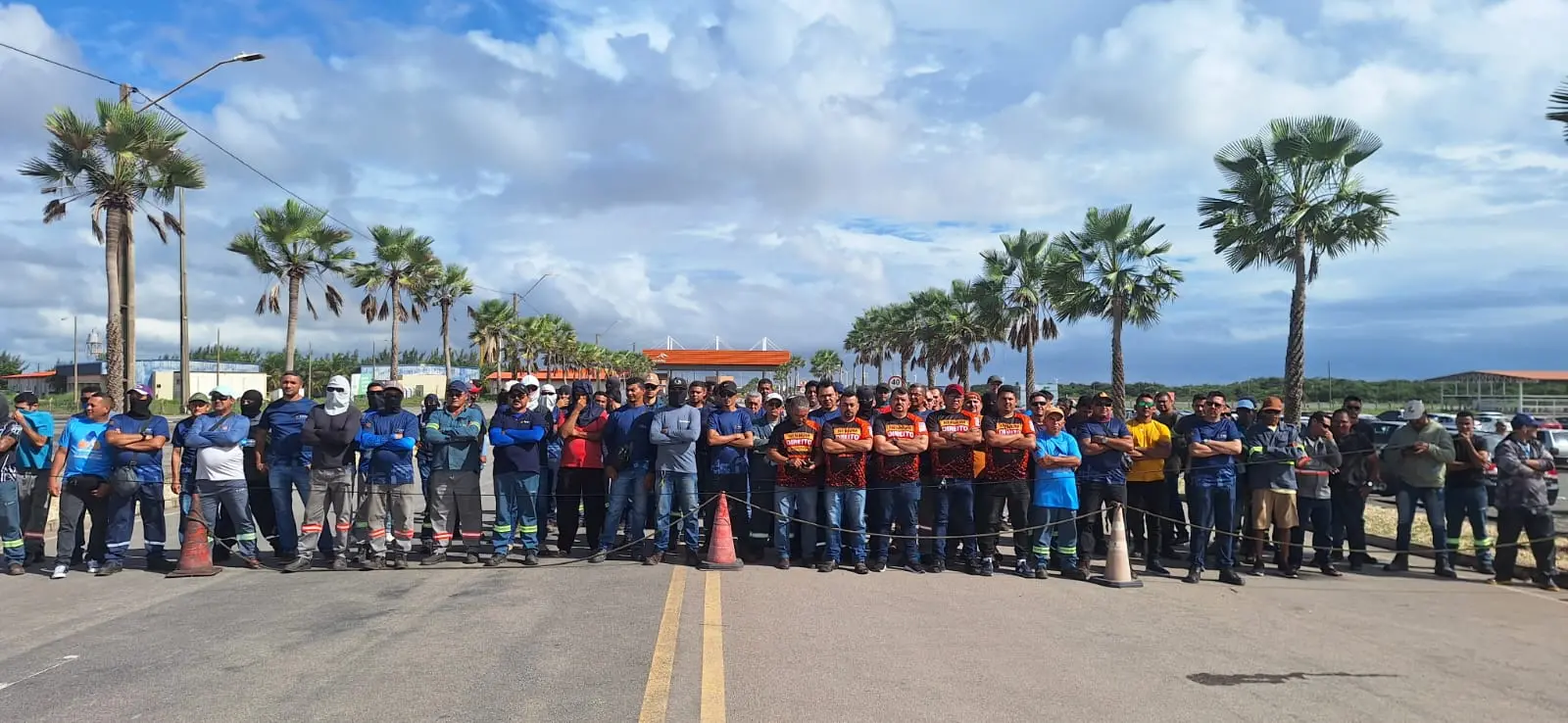 Grupo de trabalhadores reunidos em bloqueio de via em área industrial do Complexo do Pecém. Homens estão alinhados lado a lado, com braços cruzados, em frente a cones e corda de isolamento. Ao fundo, há fileiras de palmeiras, veículos estacionados e céu nublado.