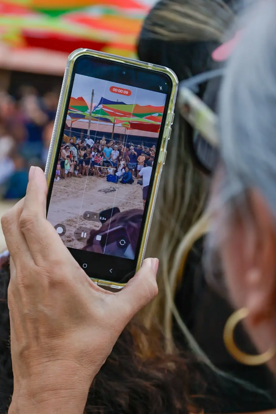 Close nas mãos de uma mulher segurando um celular que filma uma multidão em uma praia. A tela do aparelho mostra pessoas reunidas ao redor de um cercado na areia para observar tartarugas.