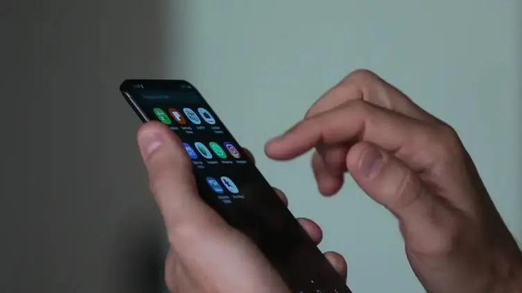 Na foto, aparece a mão esquerda de um homem branco com um celular. Na tela há alguns aplicativos instalados e a outra mão fazendo o movimento de clicar.