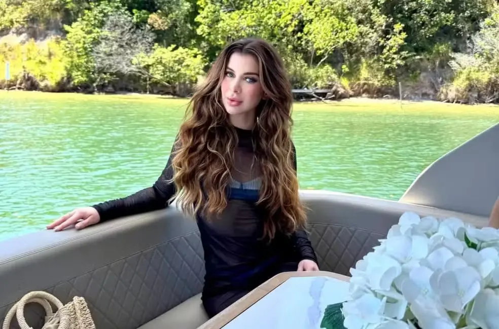 Jovem mulher com cabelo longo e cacheado, usando roupa preta, sentado em barco com vista para lago e árvores verdes ao fundo, em um ambiente natural.