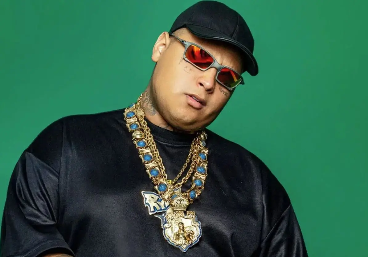 Pessoa posa em frente a um fundo verde liso, usando boné preto, óculos escuros com lentes avermelhadas, camiseta preta e vários colares dourados grandes no pescoço.