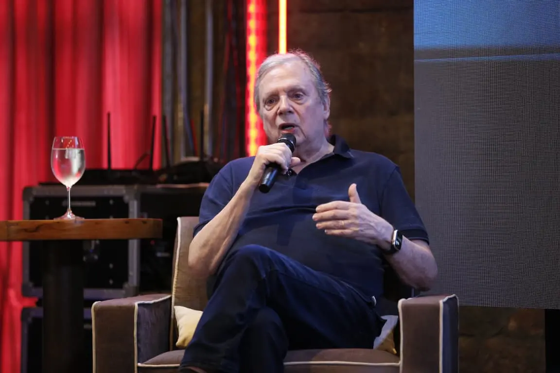 Homem idoso sentado em uma poltrona em um palco, segurando um microfone e falando. Ele veste camiseta polo escura e calça, com as pernas cruzadas. Ao lado, há uma pequena mesa com uma taça de água. O fundo é iluminado com luzes vermelhas e elementos de cenário, sugerindo um evento ou palestra.