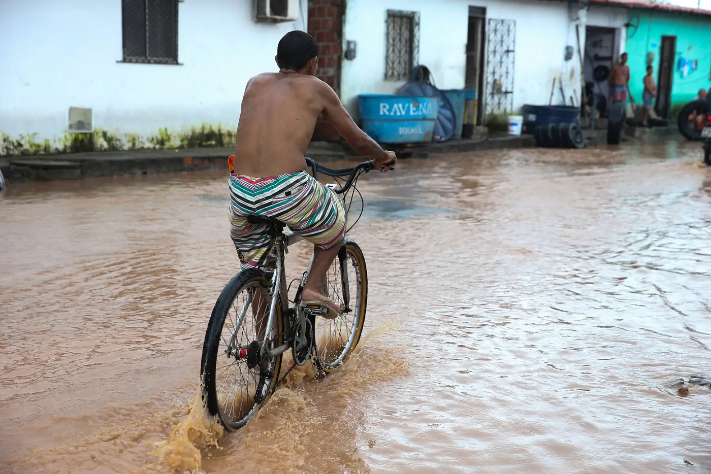 Homem negro sem camisa pedala em bicicleta em rua alagada por lama em Fortaleza. Ao fundo, há casas de muro branco e outras pessoas na calçada.