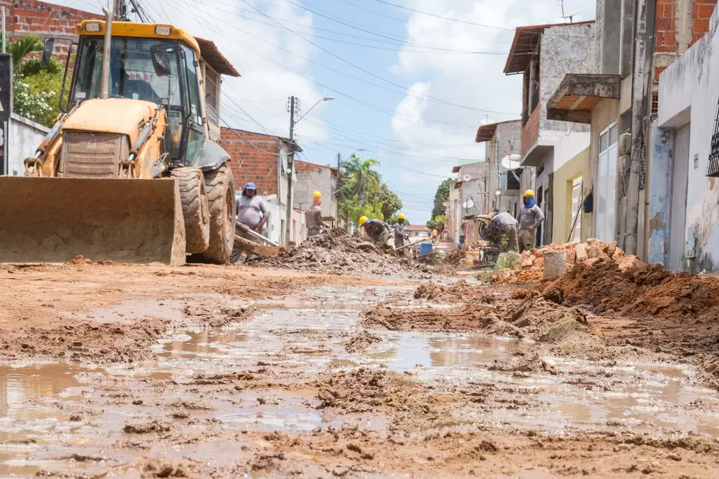 Rua enlamaçada passa por obras de saneamento, com presença de retroescavadeira e trabalhadores na periferia de Fortaleza.