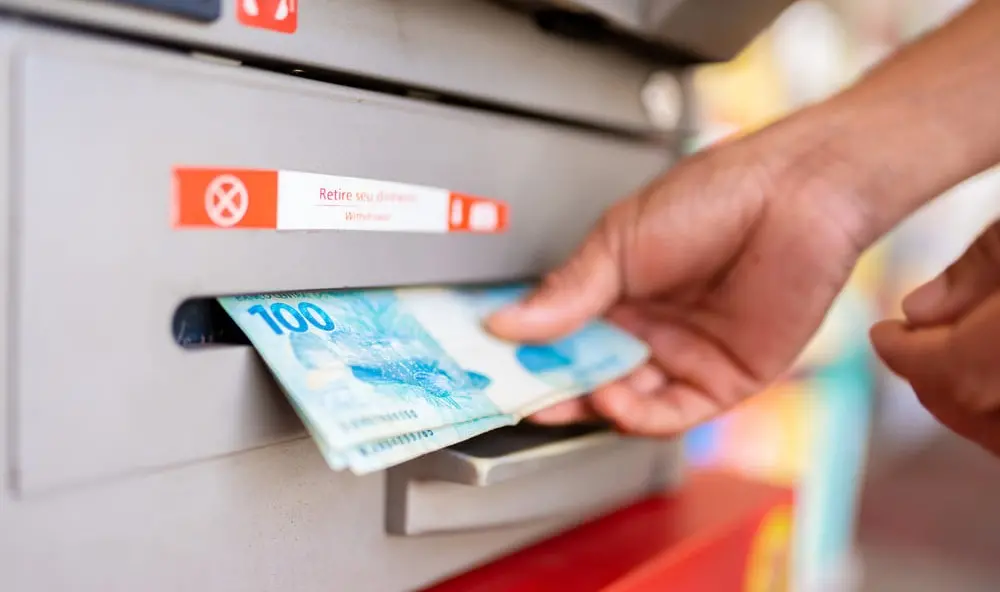 Mão masculina retira cédulas de cem reais de caixa eletrônico.