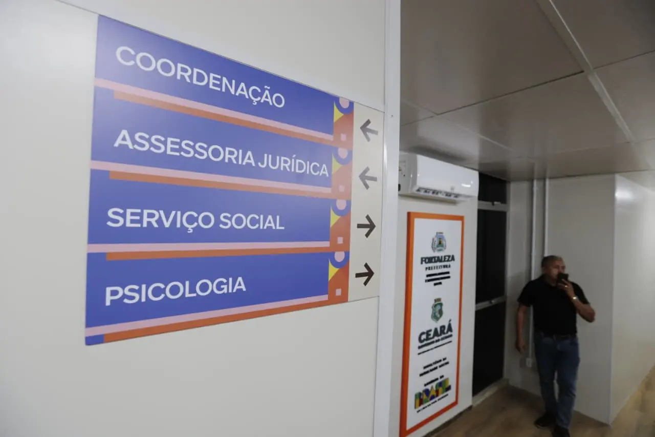 Imagem mostra sinalização de áreas de atendimento da Casa da Igualdade Racial de Fortaleza, incluindo coordenação, assessoria jurídica, serviço social e psicologia, com homem tomando nota na parede.