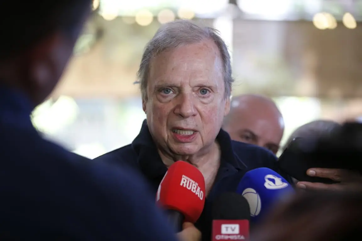 Homem idoso, de cabelos grisalhos e expressão séria, fala com a imprensa enquanto é cercado por repórteres. Vários microfones coloridos com logotipos de emissoras estão próximos ao seu rosto. Ao fundo, outras pessoas e um ambiente interno desfocado.