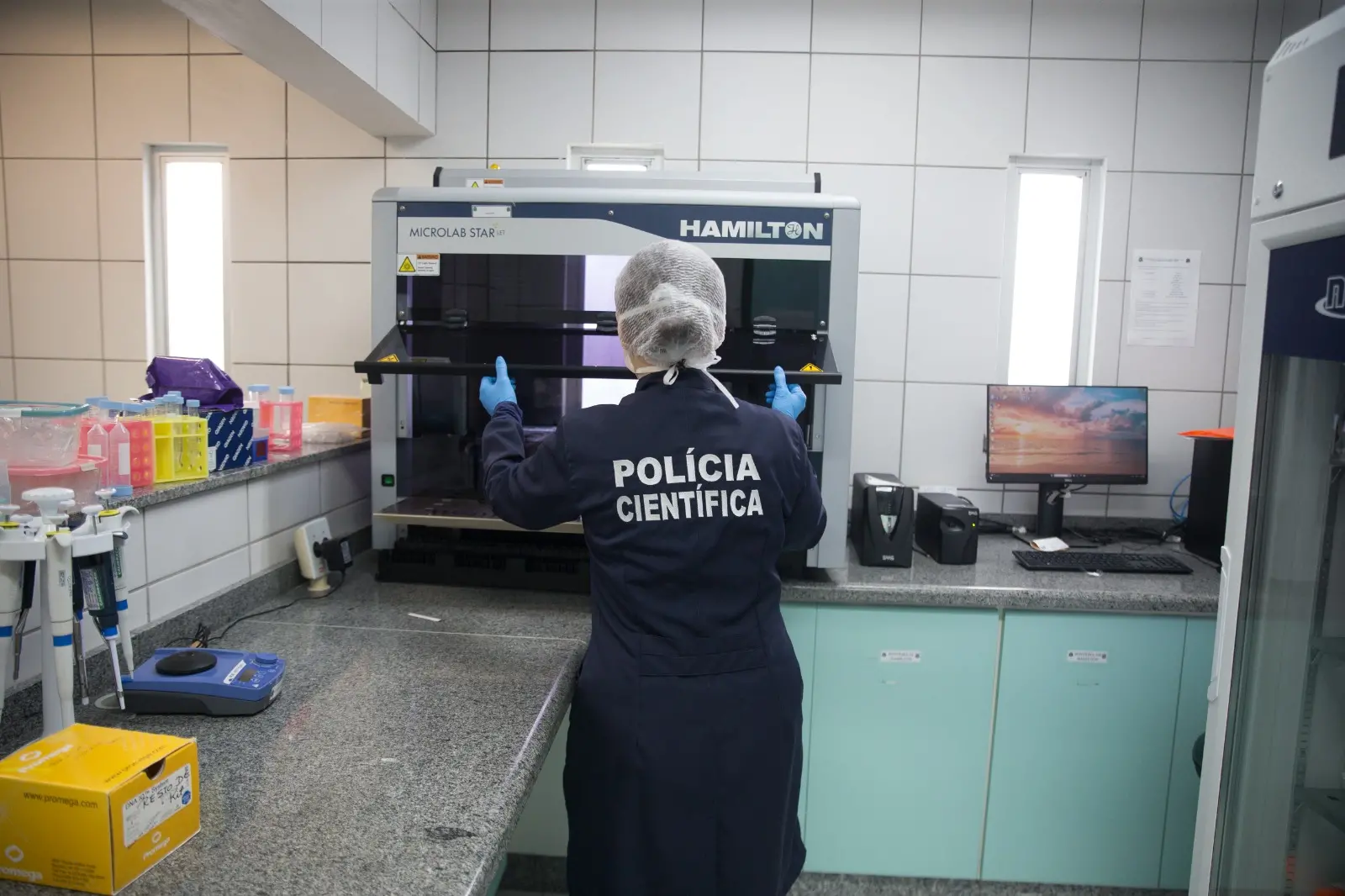Pessoa de costas, com jaleco escrito “Polícia Científica”, operando equipamento automatizado em laboratório da Pefoce, em Fortaleza.
