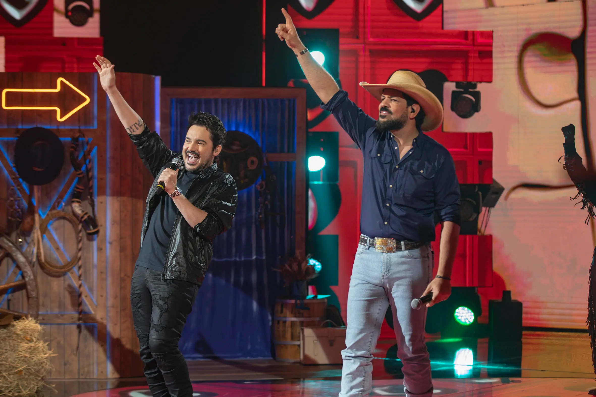 Duas pessoas cantam em um palco iluminado, segurando microfones e levantando um braço, com cenário colorido e elementos de estúdio ao fundo.