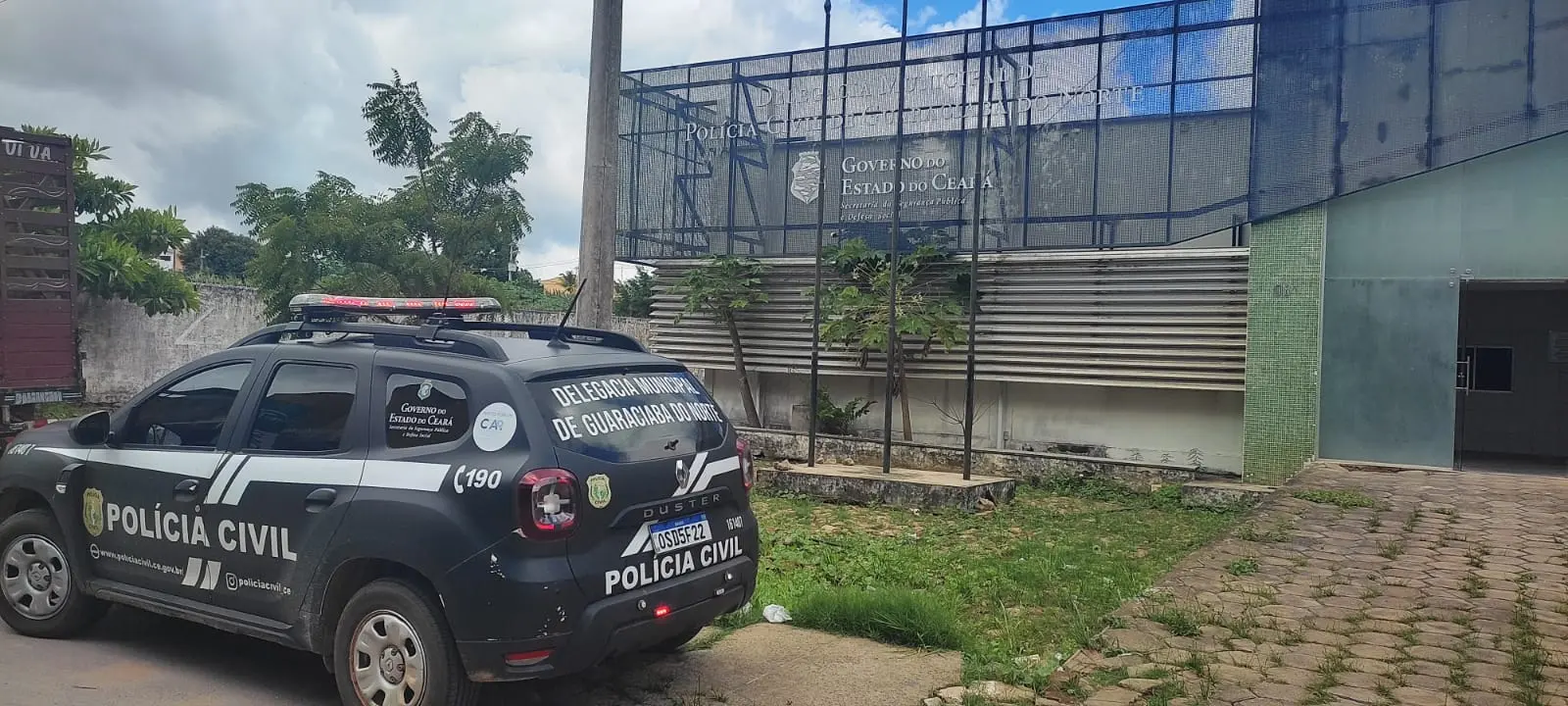 Uma viatura da Polícia Civil do Ceará aparece estacionada em frente à Delegacia Municipal de Guaraciaba do Norte, destacando os logotipos oficiais do Governo do Estado. O prédio ao fundo apresenta uma fachada moderna com grades metálicas escuras e sinalização institucional sob um céu nublado.