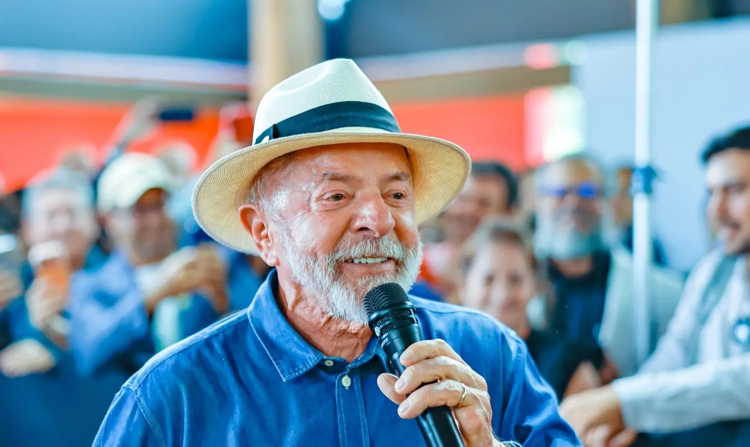 Lula é um homem branco, idoso, de cabelo e barba brancos. Na foto, ele usa uma camisa de botão azul e chapéu de palha.