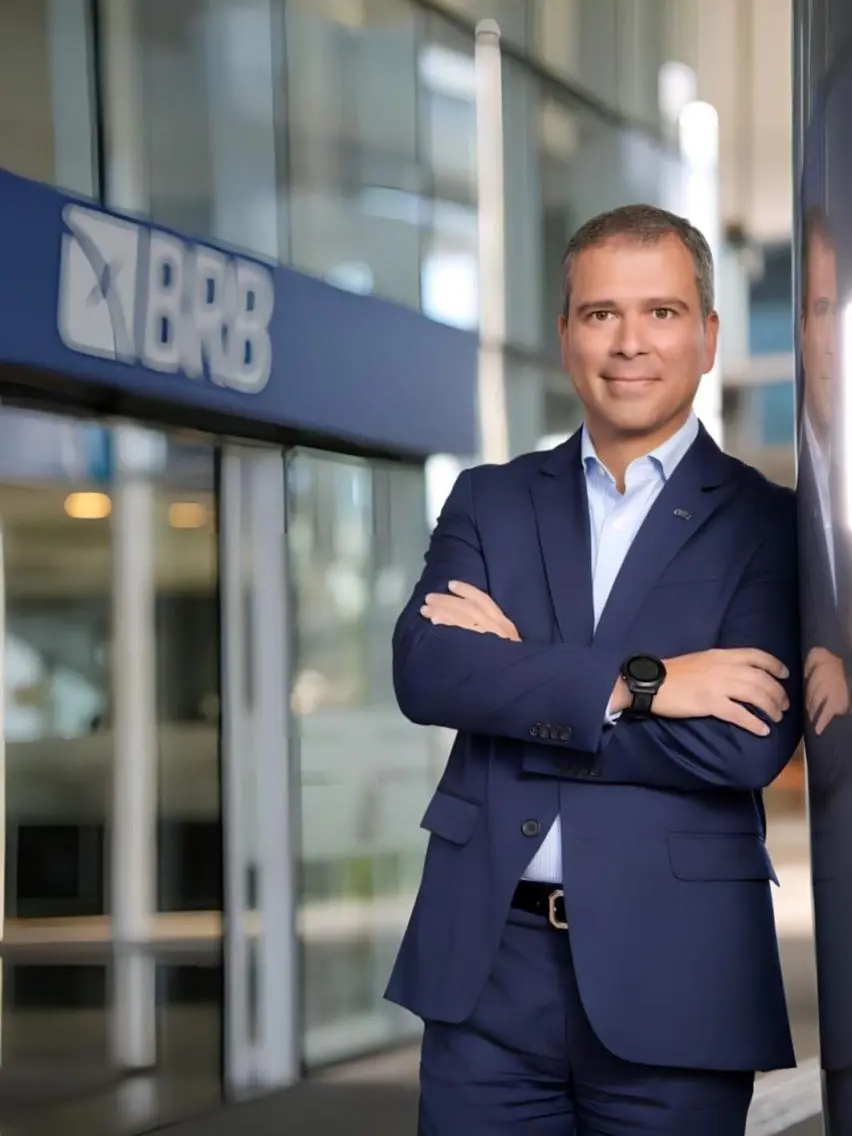 Um homem de terno azul marinho e camisa clara posa com os braços cruzados em frente a uma agência do BRB, exibindo um sorriso discreto. Ele está encostado em uma pilastra em um ambiente corporativo moderno e iluminado, com o logotipo do banco visível ao fundo.