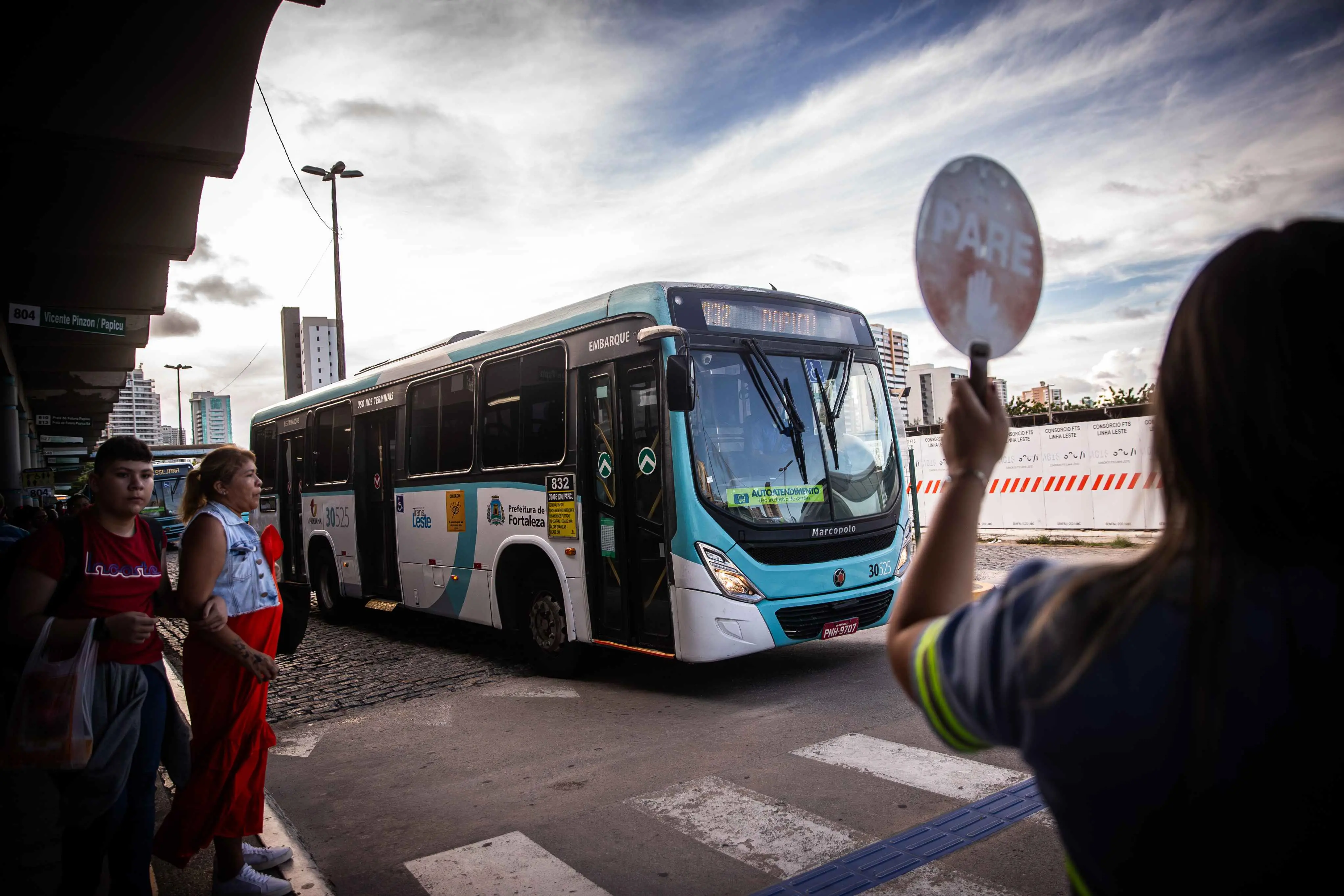 Ônibus branco e azul da Prefeitura de Fortaleza circula em terminal. Em primeiro plano, uma agente de trânsito segura uma placa de PARE enquanto pedestres aguardam para atravessar.