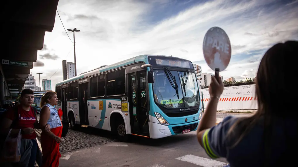 Ônibus branco e azul da Prefeitura de Fortaleza circula em terminal. Em primeiro plano, uma agente de trânsito segura uma placa de PARE enquanto pedestres aguardam para atravessar.