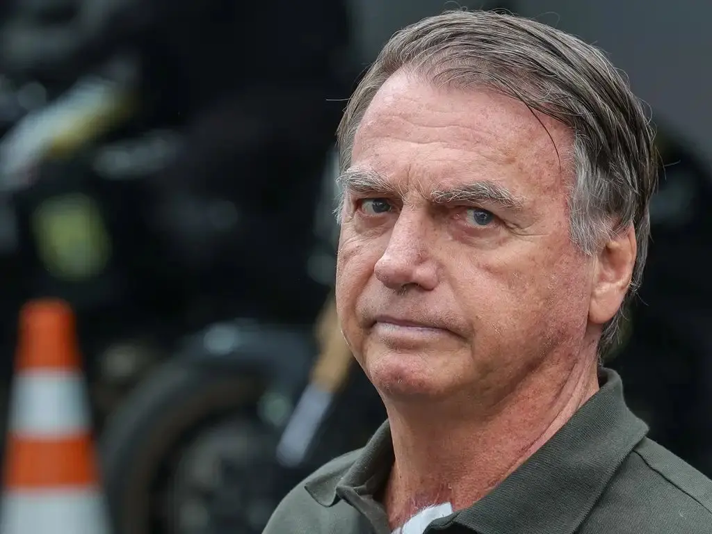 Jair Bolsonaro com expressão séria, cabelos grisalhos e camisa polo cinza, na rua com fundo de trânsito, incluindo um cone de trânsito.