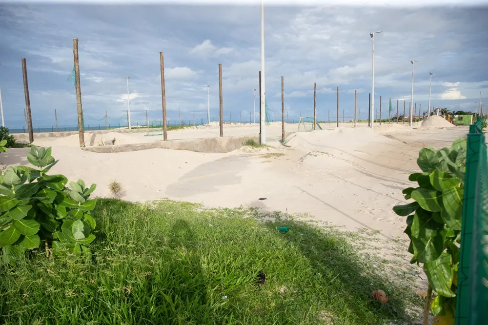 Quadras esportivas de areia com postes de madeira e redes sob um céu nublado; grama verde e plantas em primeiro plano.