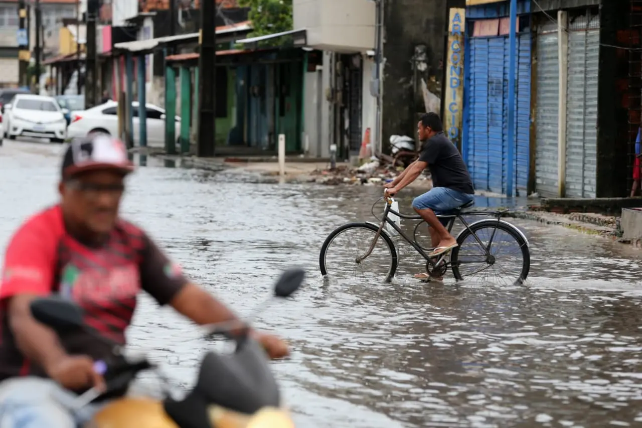 esta imagem mostra homens circulando em áreas alagadas por água da chuva. um de moto e outro dd bicicleta.