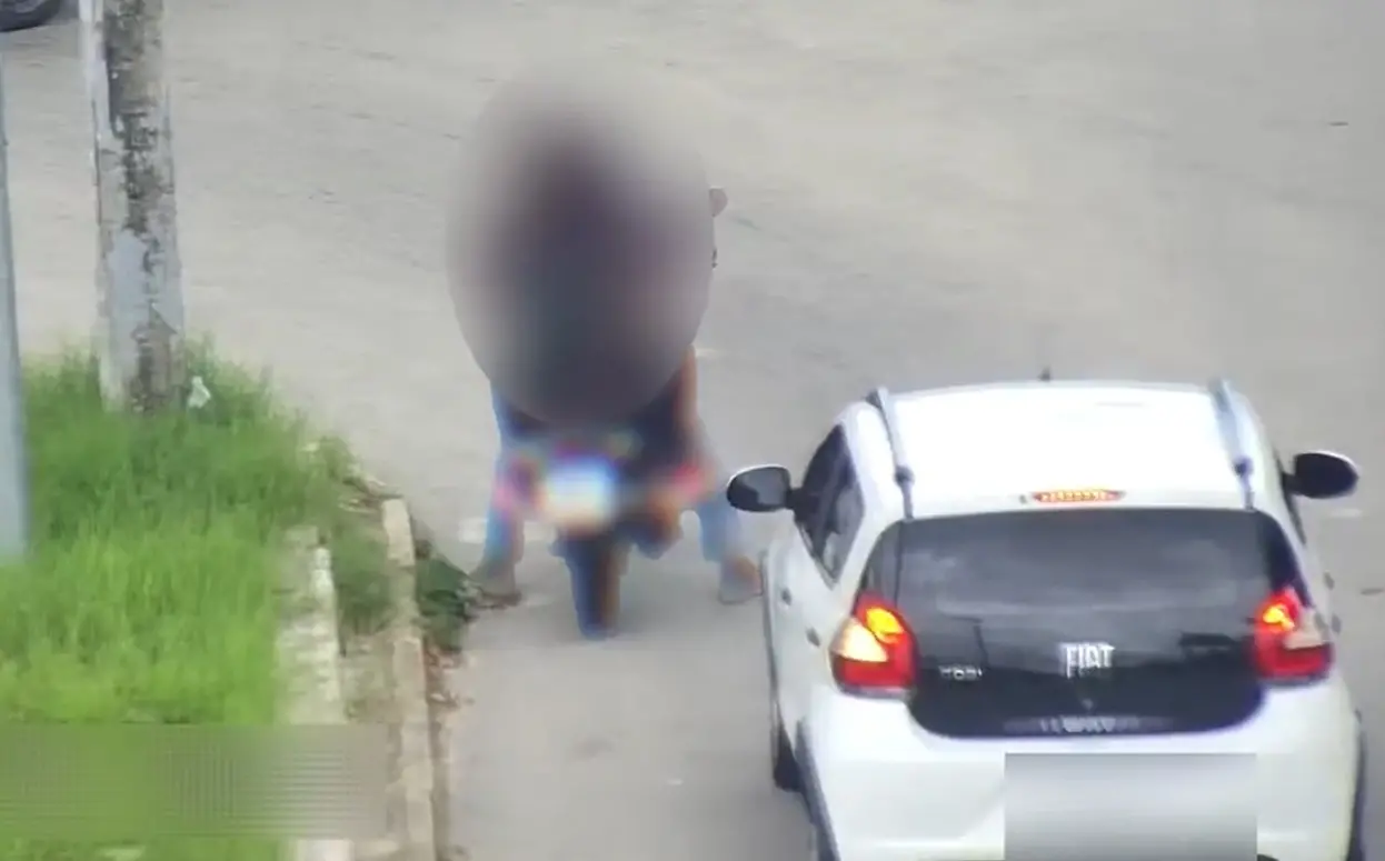 Em uma imagem com tarjas, é possível ver o suspeito transportando uma passageira. A motocicleta está parada em um semáforo.