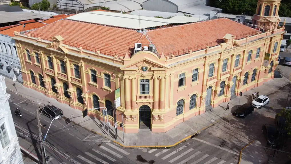 Imagem aérea de um prédio histórico de arquitetura clássica colorido em tons de creme e laranja, localizado na orla de uma cidade costeira com mar e céu azul ao fundo.