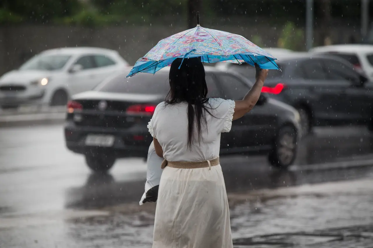 Uma mulher de costas segura um guarda-chuva colorido enquanto caminha sob chuva intensa, com o asfalto molhado refletindo as luzes dos carros ao fundo. O cenário urbano destaca o movimento dos veículos embaçados pela precipitação, transmitindo uma atmosfera cotidiana da cidade.