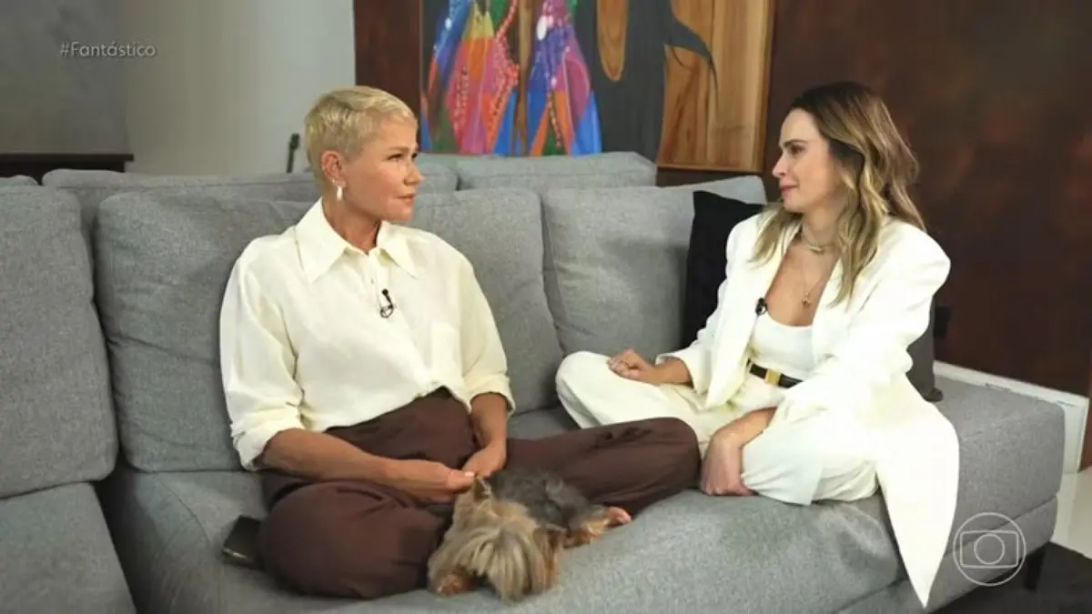 Duas pessoas adultas posam juntas sorrindo, em ambiente interno iluminado. Uma veste roupa clara e a outra usa roupa escura. As duas estão próximas, com os braços tocando os ombros, diante de um fundo neutro.