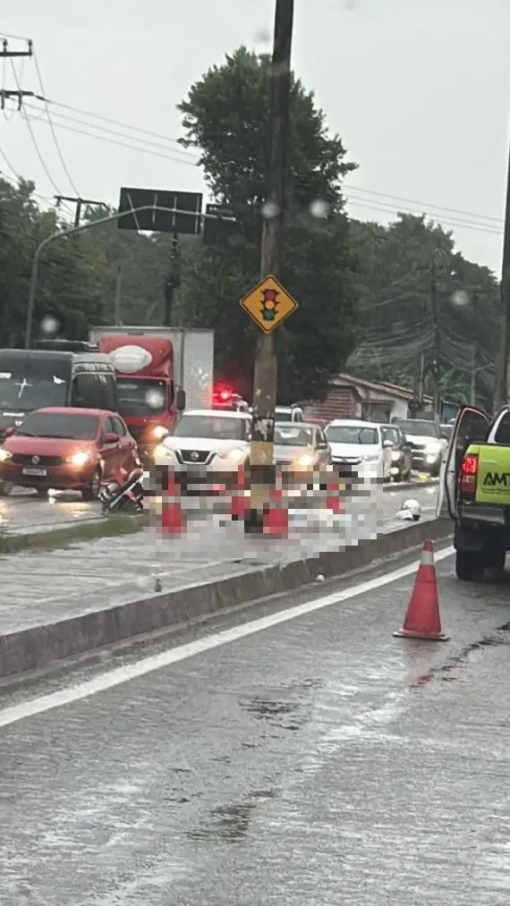 Veículos trafegam por uma via molhada sob chuva, onde uma área está isolada por cones alaranjados devido a um acidente envolvendo uma motocicleta caída. Ao fundo, o trânsito flui lentamente entre postes de iluminação e sinalizações viárias.