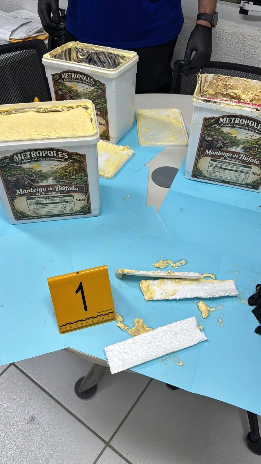 Latas abertas de manteiga marca Metrópoles com substância amarela semelhante a gordura vegetal sobre mesa azul, resíduos espalhados, fragmentos do material e marcador numerado em ambiente técnico. Dentro das latas havia maconha escondida.