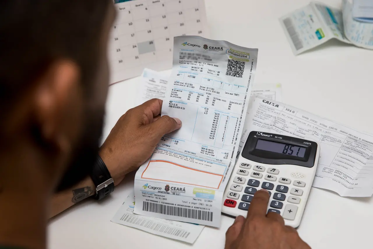Um homem analisa uma conta de água da Cagece enquanto utiliza uma calculadora sobre uma mesa repleta de boletos e um calendário ao fundo. A imagem, capturada em close-over-the-shoulder, destaca o planejamento financeiro doméstico através do manuseio de documentos de cobrança e cálculos manuais.