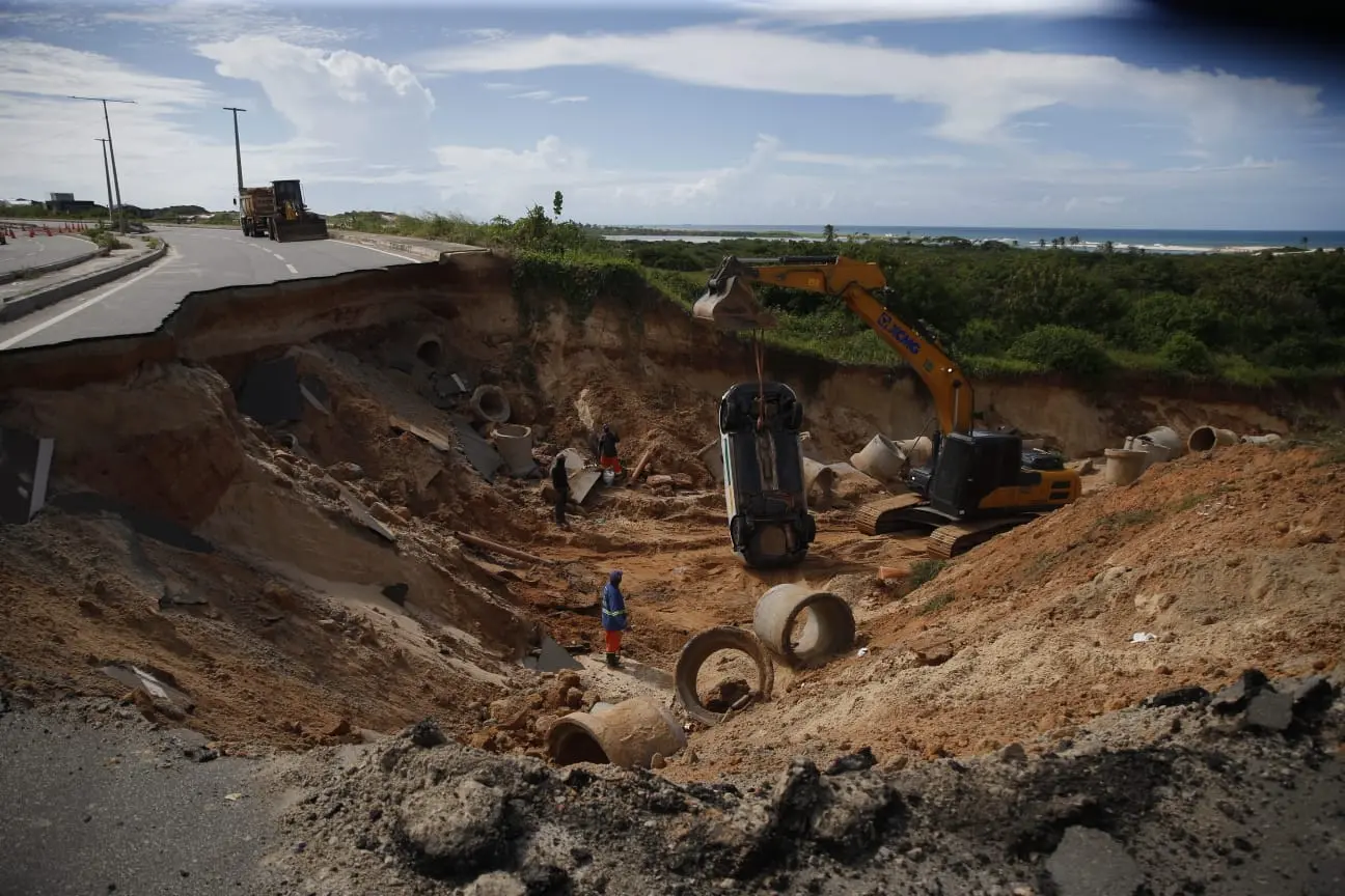 Rodovia parcialmente destruída por uma grande cratera em área costeira; escavadeira remove um carro virado enquanto trabalhadores atuam entre entulhos e manilhas expostas, com o mar e vegetação ao fundo sob céu parcialmente nublado.