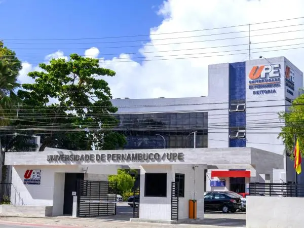 Fachada da Reitoria da Universidade de Pernambuco (UPE), composta por um pórtico de entrada em tons claros e um edifício moderno ao fundo com detalhes em azul. A composição destaca o logotipo da instituição sob um céu ensolarado com nuvens, transmitindo a sobriedade e a importância do ambiente acadêmico.