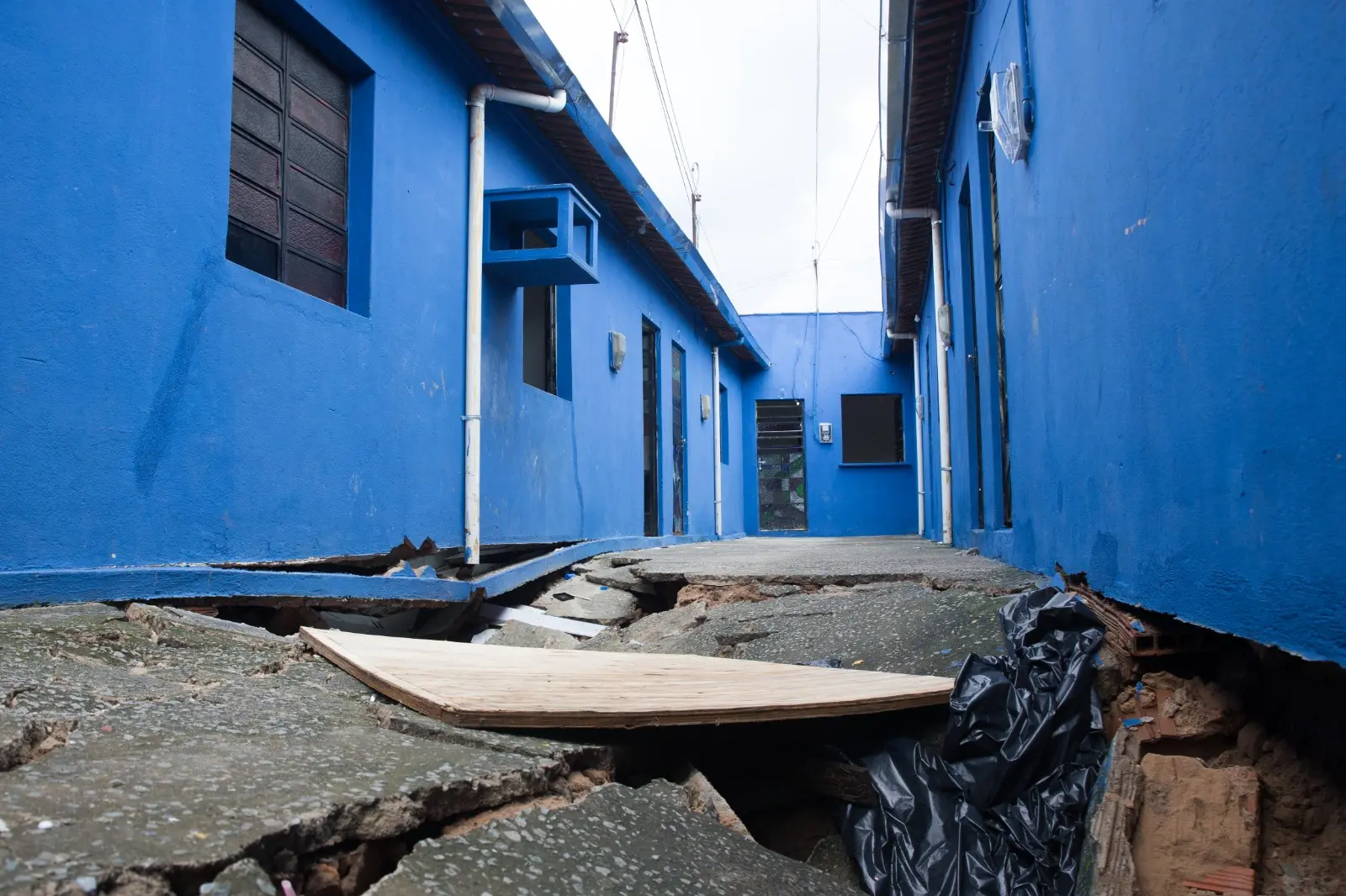 Piso de concreto totalmente fragmentado e cedido no corredor estreito de uma vila com paredes azuis vibrantes em Fortaleza. O desabamento do solo expõe o interior da fundação, com pedaços de madeira e sacos plásticos sobre os escombros.