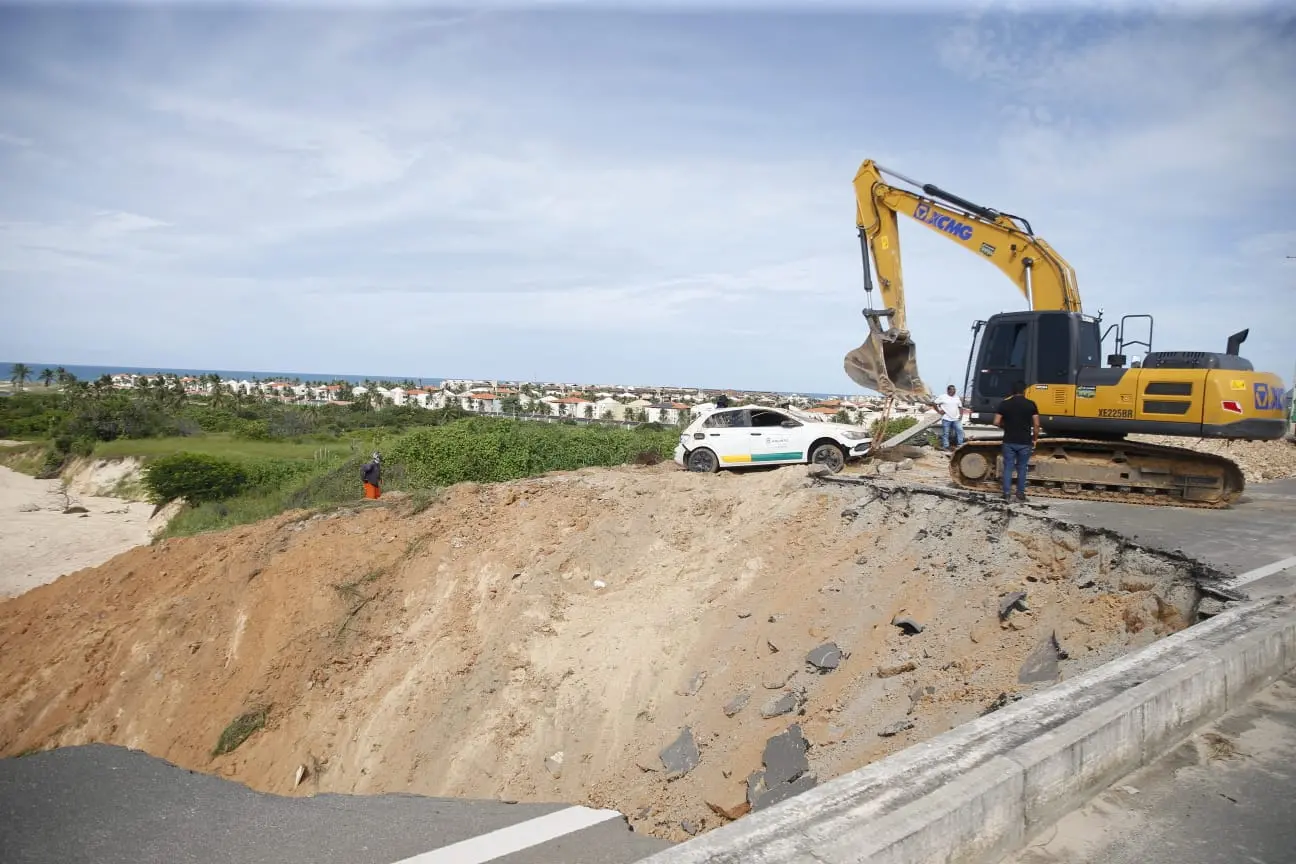 Escavadeira amarela opera na borda de uma grande cratera em uma estrada asfaltada de Aquiraz, onde parte da pista desmoronou. Ao fundo, um carro branco está parado perigosamente próximo à erosão, com vista para uma área residencial e o mar.