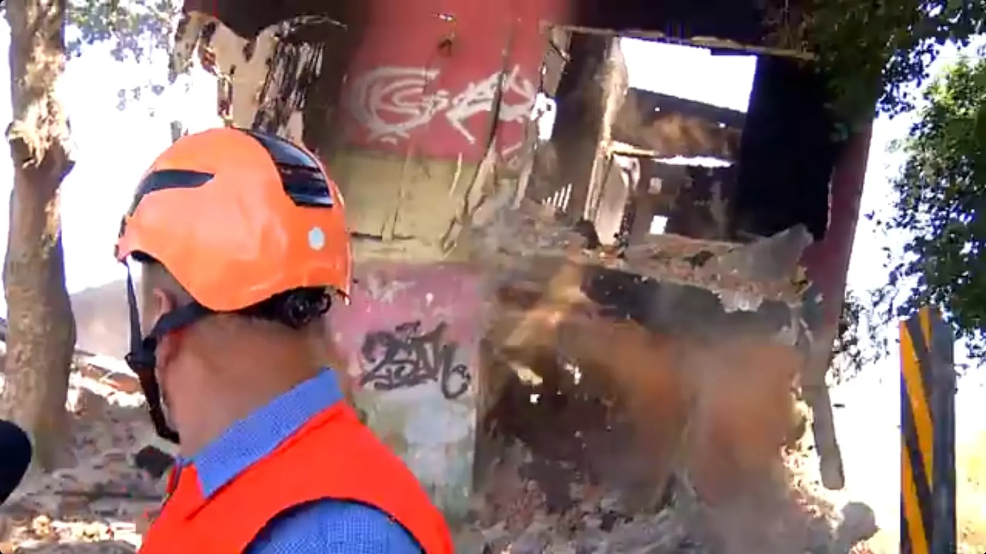 Um profissional da Defesa Civil com capacete laranja e colete reflexivo observa os escombros de um imóvel desabado com paredes pichadas. A cena captura o momento em que uma nuvem de poeira se levanta entre as ruínas e árvores sob a luz forte do dia.