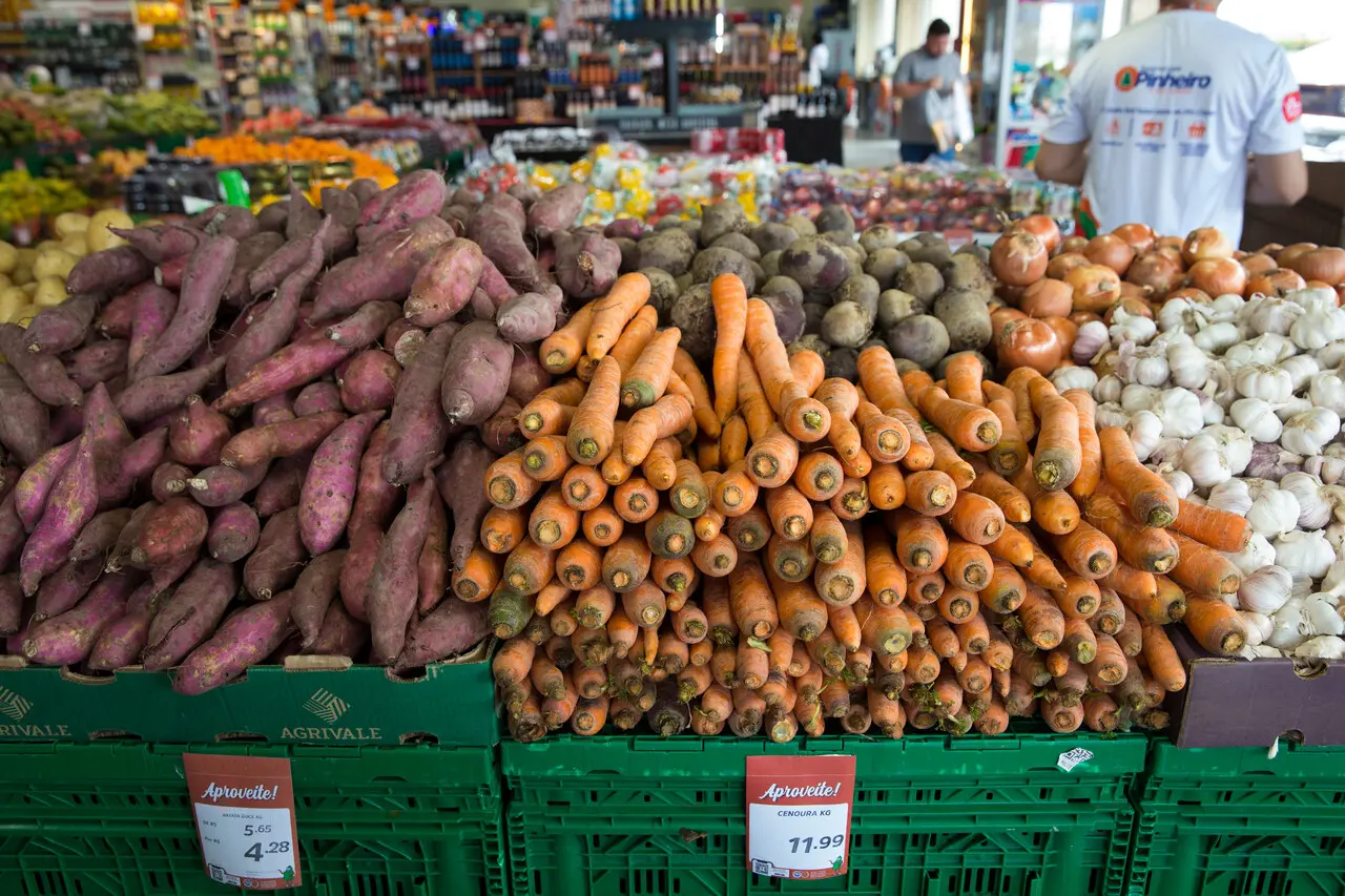 Bancadas de um supermercado exibem pilhas organizadas de batata-doce, cenouras, beterrabas, cebolas e alho em caixas plásticas verdes com etiquetas de preços em destaque. Ao fundo, o corredor da loja aparece levemente desfocado com clientes e funcionários circulando entre as prateleiras de hortifrúti.