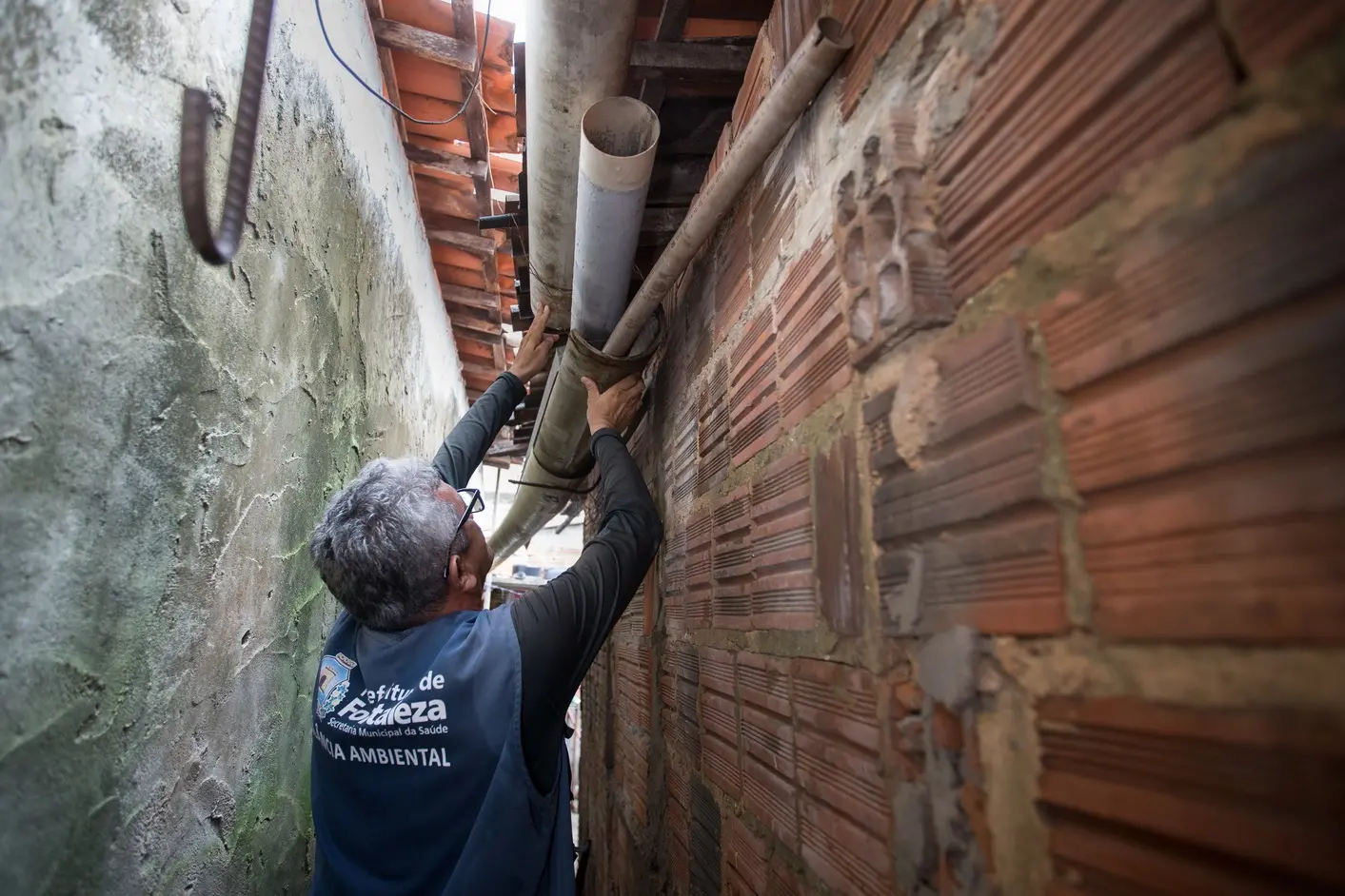Um agente de vigilância ambiental, visto de costas, inspeciona calhas de chuva no telhado de uma casa em um beco estreito. Ele utiliza as mãos para verificar possíveis acúmulos de água parada que possam servir de criadouro para o mosquito Aedes aegypti.