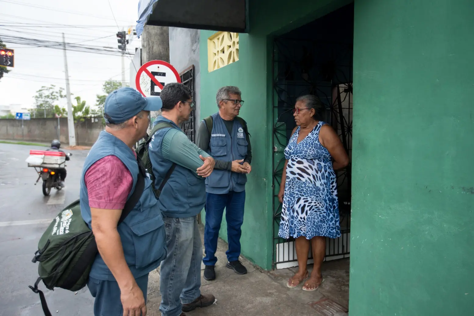 Três agentes de endemias da Prefeitura de Fortaleza, vestindo coletes azuis, conversam com uma moradora à porta de sua residência. A ação educativa ocorre em uma calçada próxima a um cruzamento, com o objetivo de orientar sobre a prevenção da dengue.