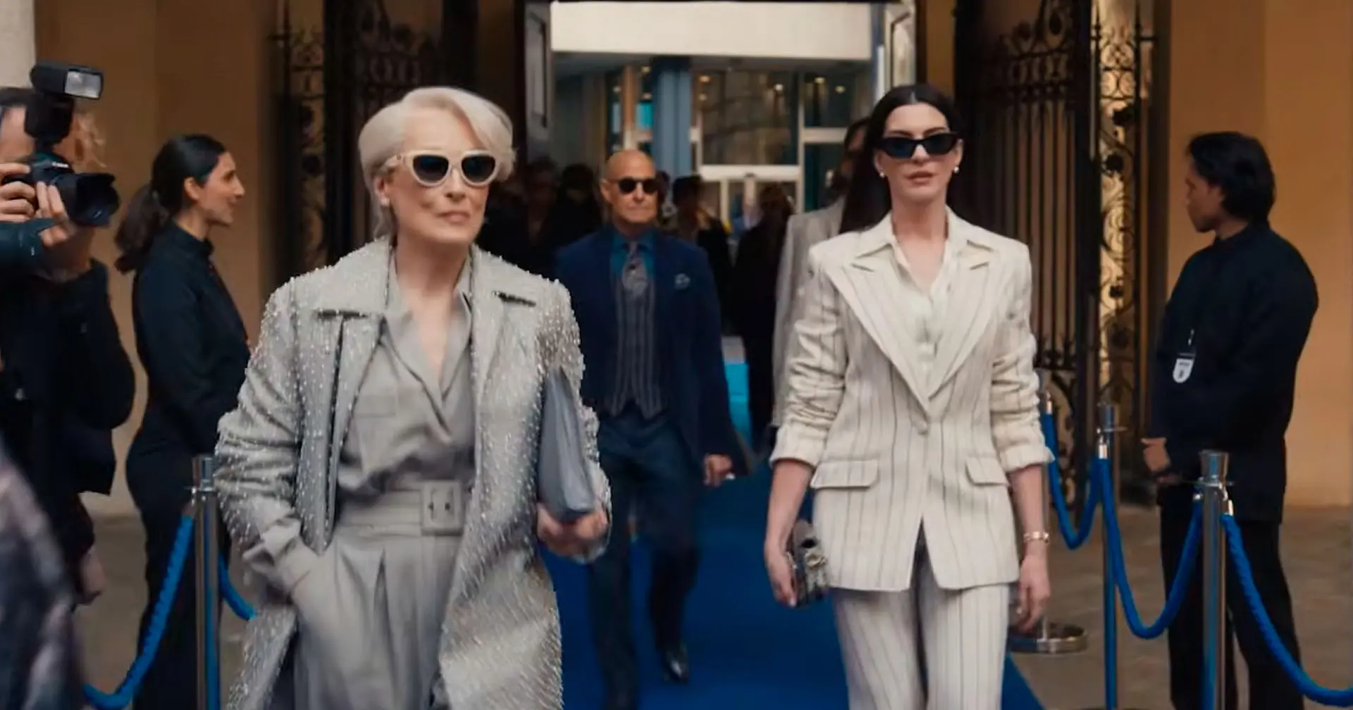 Três pessoas caminhando em um corredor interno iluminado, vestidas com roupas sociais em tons neutros, carregando bolsas e pastas, com vitrines e pessoas desfocadas ao fundo.