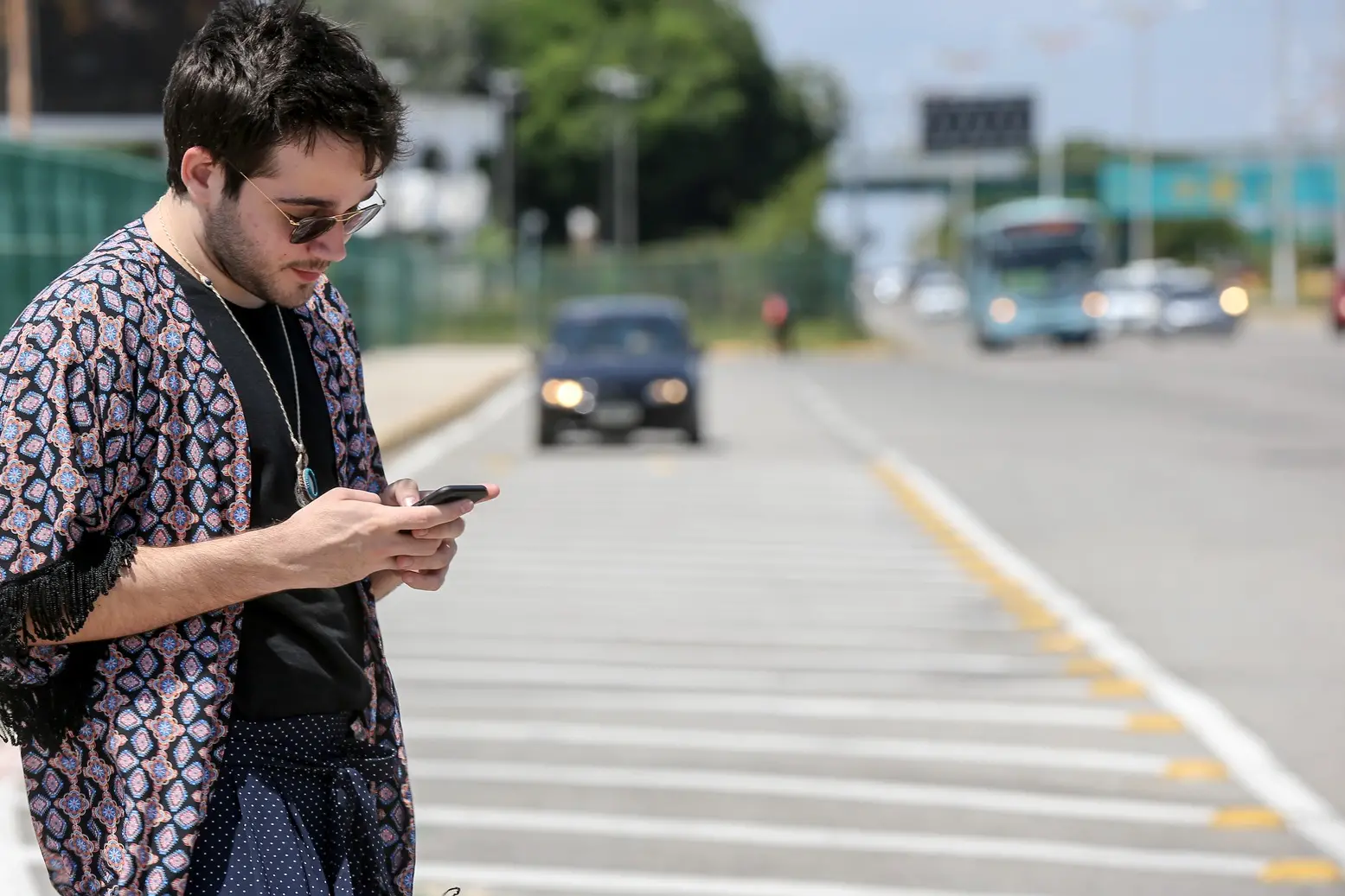 Homem branco ao celular solicitando um uber. Ao fundo aparece um carro preto.