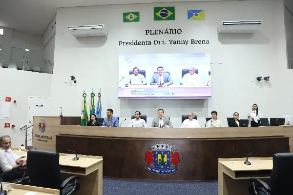 Plenário de uma câmara municipal com mesa diretora ao fundo, onde várias pessoas estão sentadas em posição de reunião. À frente, há mesas com cadeiras vazias. Na parede, acima do painel, lê-se “PLENÁRIO Presidenta Dra. Yanny Brena”, acompanhado de bandeiras do Brasil, do estado e do município. Um telão exibe a imagem ampliada dos participantes da mesa. O ambiente é bem iluminado, com paredes claras e ar-condicionado instalado.