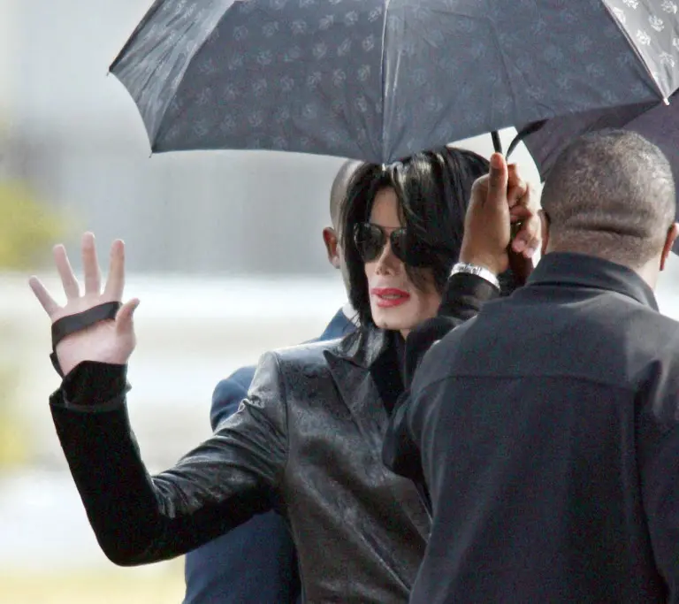 Michael Jackson com cabelo preto, roupas escuras, usando óculos escuros, segurando um guarda-chuva preto, acenando com a mão ao público em evento público
